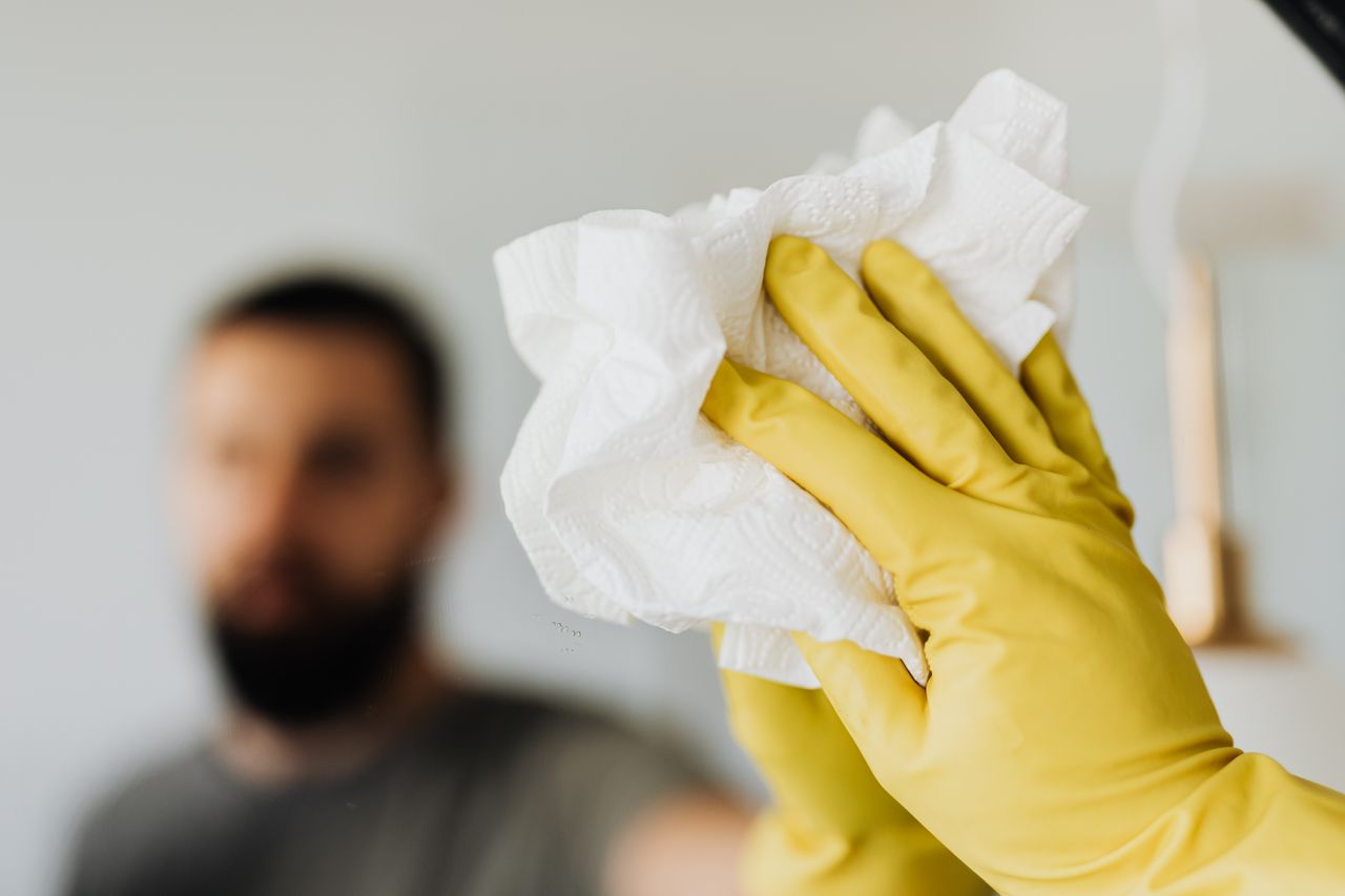 Homme barbu non identifiable, équipé de gants en latex jaunes, essuyant le miroir avec une serviette dans la salle de bain à la maison